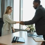 Two business professionals shaking hands across a desk in a bright office.